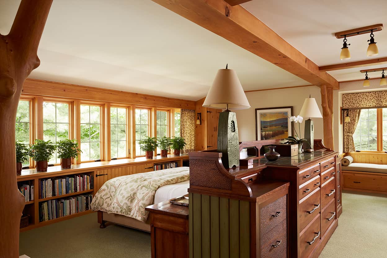 Master bedroom with natural wood woodwork, pendant lighting, muted blue-green carpet, low bookshelves under the windows, and a television that lowers into cabinetry when not wanted
