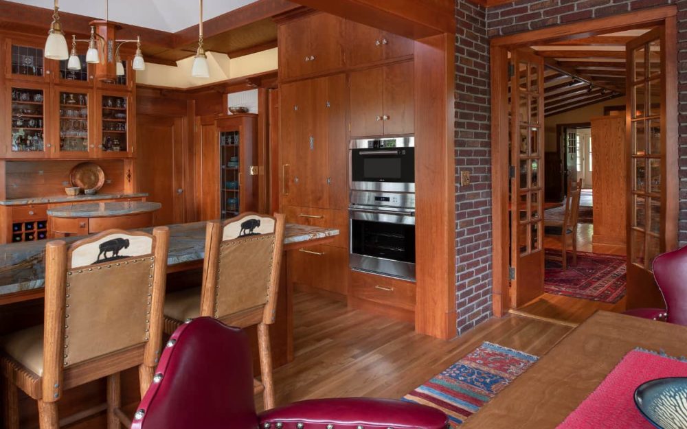 The dining room table is paired with a mix of wood straight chairs and red leather arm chairs, and the stools at the counter are tan leather with a buffalo design at the top. The dining room's brick wall opens up into the rest of the house, a glimpse of beamed ceilings, panelling, and plenty of light.