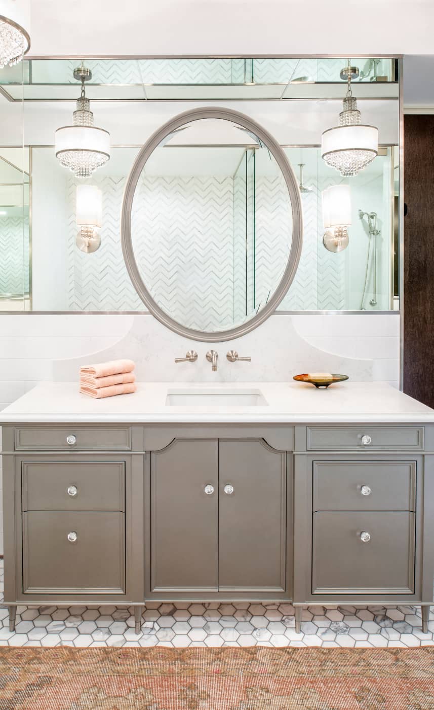 Start with an elegantly simple gray vanity with a white quartz counter atop a gray and white marble hex tiled floor. Add a gray-framed oval mirror mounted on a tall strip of mirrors framed by white stone, all under a mirrored ceiling. Finish off with crystal chandeliers. The result is a dazzling symphony of gray, white, and glass.
